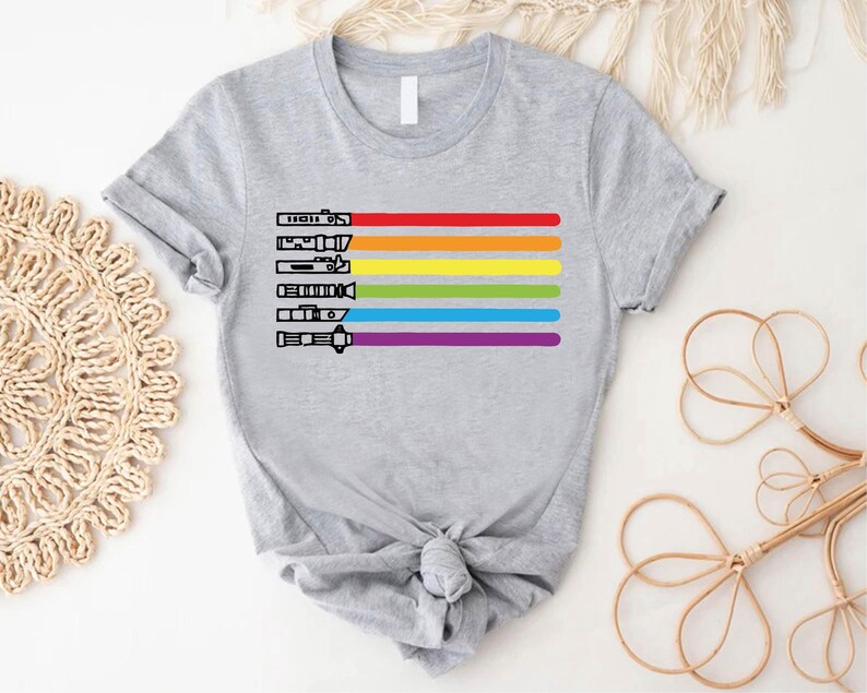 Puede incluir: Una camiseta gris con un dise&ntilde;o de arco&iacute;ris y siluetas de sables de luz negros. El arco&iacute;ris est&aacute; formado por rayas rojas, naranjas, amarillas, verdes, azules, &iacute;ndigo y violetas.