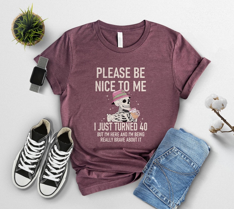 May include: Burgundy t-shirt with the text "PLEASE BE NICE TO ME I JUST TURNED 40". The graphic features a skeleton wearing a hat and holding a drink. The shirt is paired with blue jeans and black sneakers.