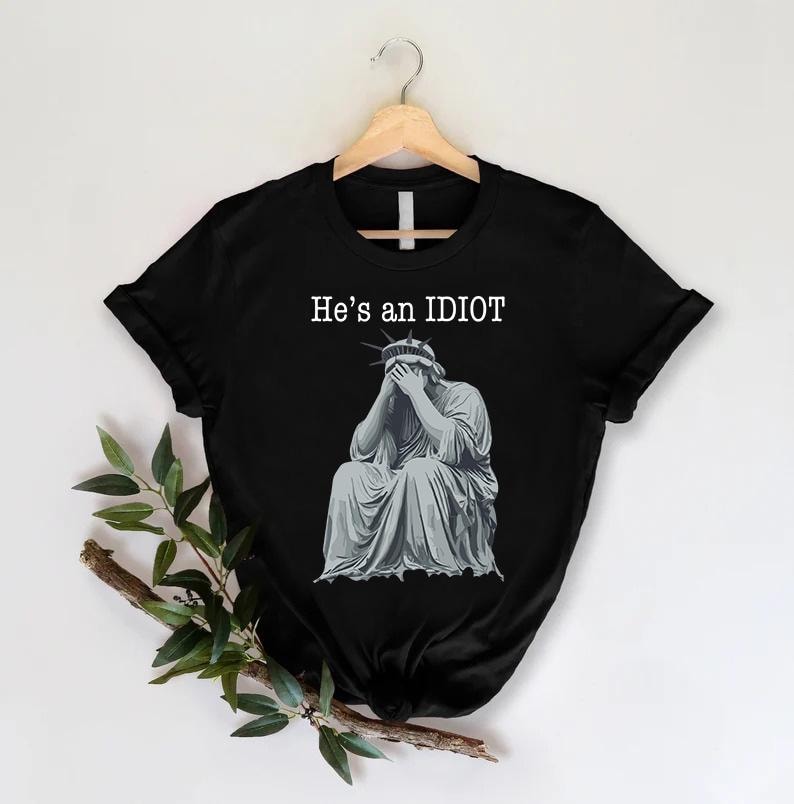 May include: Black t-shirt featuring the text "He's an IDIOT" above a grey illustration of the Statue of Liberty, head in hands. The shirt is displayed on a wooden hanger.