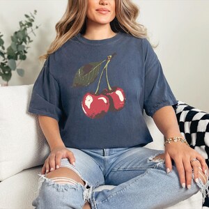 May include: Navy blue t-shirt featuring a red cherry graphic with a green leaf. The shirt has short sleeves and a relaxed fit. The model is wearing light blue ripped jeans.