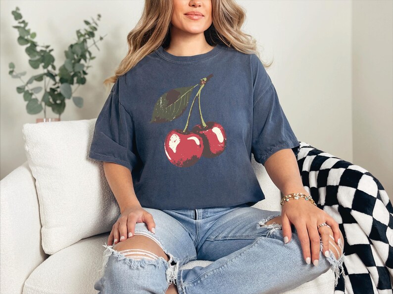 May include: Navy blue t-shirt featuring a red cherry graphic with a green leaf. The shirt has short sleeves and a relaxed fit. The model is wearing light blue ripped jeans.