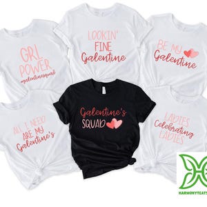 May include: White and black t-shirts with pink text and graphics. The shirts have phrases like "GRL POWER," "LOOKIN' FINE Galentine," and "Galentine's SQUAD." The shirts are for a Galentine's Day theme.