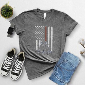 Puede incluir: Camiseta gris con un dise&ntilde;o desgastado de la bandera estadounidense, una silueta de personaje y un sable de luz rojo. La camiseta se combina con zapatillas negras, un reloj inteligente y vaqueros azules.