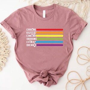 Puede incluir: Una camiseta malva con un dise&ntilde;o de arco&iacute;ris que presenta una fila de sables de luz. Los sables de luz est&aacute;n delineados en blanco y el arco&iacute;ris est&aacute; formado por rayas rojas, naranjas, amarillas, verdes, azules, &iacute;ndigo y violetas.