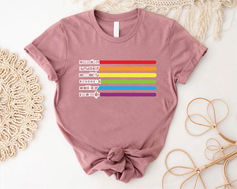 Puede incluir: Una camiseta malva con un dise&ntilde;o de arco&iacute;ris que presenta una fila de sables de luz. Los sables de luz est&aacute;n delineados en blanco y el arco&iacute;ris est&aacute; formado por rayas rojas, naranjas, amarillas, verdes, azules, &iacute;ndigo y violetas.