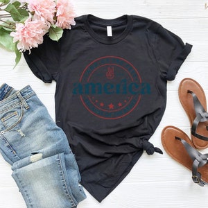 May include: Black t-shirt with a red, white, and blue graphic that says "Land of the Free, America, Because of the Brave". The graphic features a peace sign and stars.