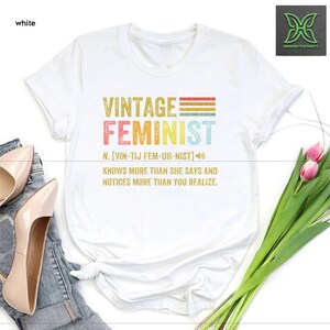 May include: White t-shirt with a vintage feminist graphic design. The graphic features the word "Feminist" in rainbow colors with a definition and a vintage style font.