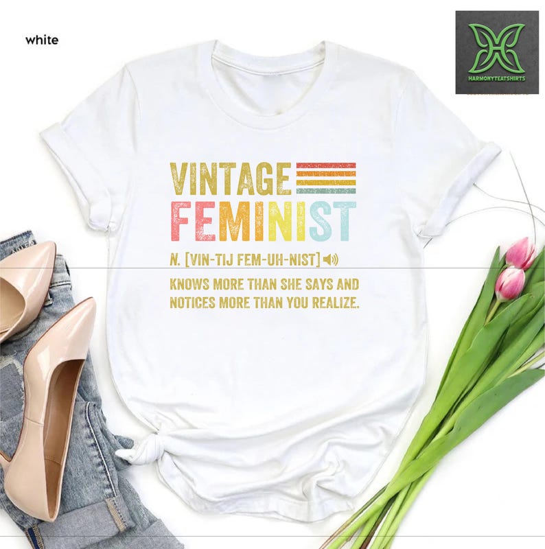 May include: White t-shirt with a vintage feminist graphic design. The graphic features the word "Feminist" in rainbow colors with a definition and a vintage style font.