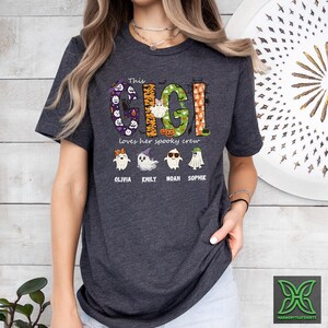 Op de afbeelding: Donkergrijs t-shirt met de tekst "This Gigi loves her spooky crew". Het ontwerp bevat het woord "Gigi" in een Halloween-thema lettertype, samen met illustraties van geesten en de namen Olivia, Emily, Noah en Sophie.
