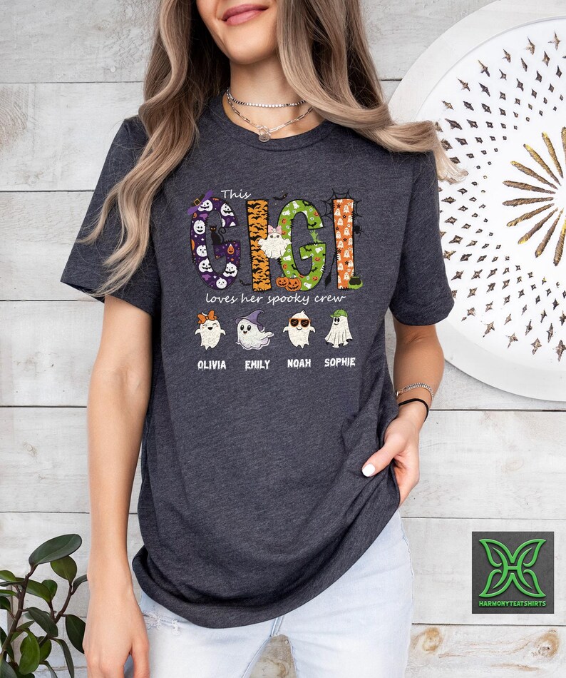 Op de afbeelding: Donkergrijs t-shirt met de tekst "This Gigi loves her spooky crew". Het ontwerp bevat het woord "Gigi" in een Halloween-thema lettertype, samen met illustraties van geesten en de namen Olivia, Emily, Noah en Sophie.