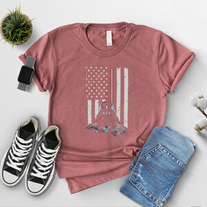 Puede incluir: Camiseta rosa polvorienta con un dise&ntilde;o de bandera estadounidense desgastada y una silueta de figura. La imagen incluye vaqueros azules, zapatillas altas negras, un reloj inteligente y una peque&ntilde;a planta en maceta. La camiseta es el foco.