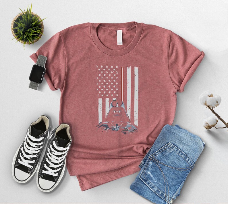 Puede incluir: Camiseta rosa polvorienta con un dise&ntilde;o de bandera estadounidense desgastada y una silueta de figura. La imagen incluye vaqueros azules, zapatillas altas negras, un reloj inteligente y una peque&ntilde;a planta en maceta. La camiseta es el foco.