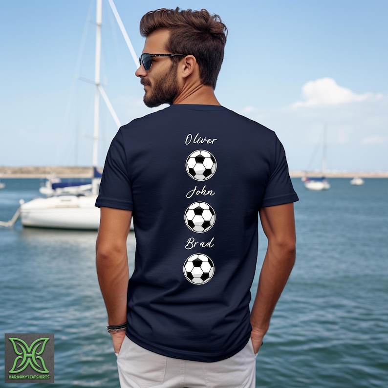 Peut inclure: T-shirt bleu marine avec trois motifs de ballon de football et les noms Oliver, John et Brad imprim&eacute;s au-dessus de chaque ballon. La personne porte des lunettes de soleil et se tient pr&egrave;s de l'eau.