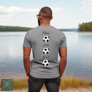 Peut inclure: Un t-shirt gris avec les noms Oliver, John et Brad, chacun au-dessus d'un ballon de football. La personne portant le t-shirt se tient devant un lac. Le logo Harmony Tee Shirts est visible en bas &agrave; gauche.