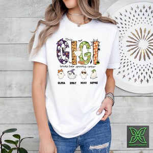 Op de afbeelding: Wit T-shirt met een Halloween-thema. Het shirt toont de tekst "This Gigi loves her spooky crew" in een kleurrijk, decoratief lettertype. Onder de tekst staan vier cartoongeesten met de namen: Olivia, Emily, Noah en Sophie.
