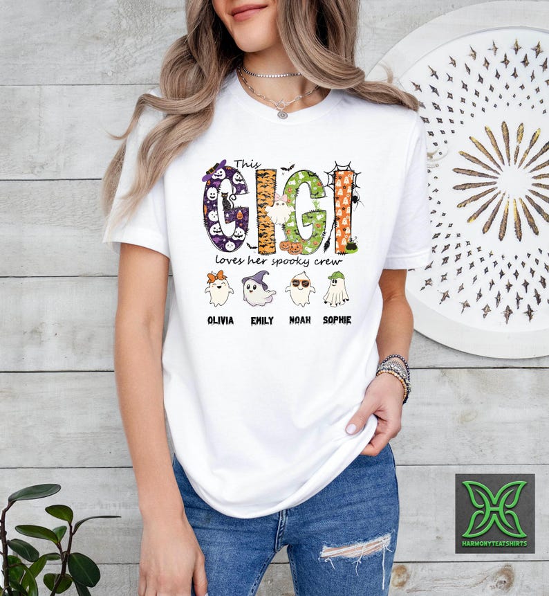 Op de afbeelding: Wit T-shirt met een Halloween-thema. Het shirt toont de tekst "This Gigi loves her spooky crew" in een kleurrijk, decoratief lettertype. Onder de tekst staan vier cartoongeesten met de namen: Olivia, Emily, Noah en Sophie.
