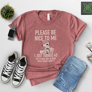 May include: A dusty rose t-shirt with the text "PLEASE BE NICE TO ME I JUST TURNED 40". The graphic shows a skeleton wearing a hat and holding a drink. The shirt is paired with blue jeans and black sneakers.