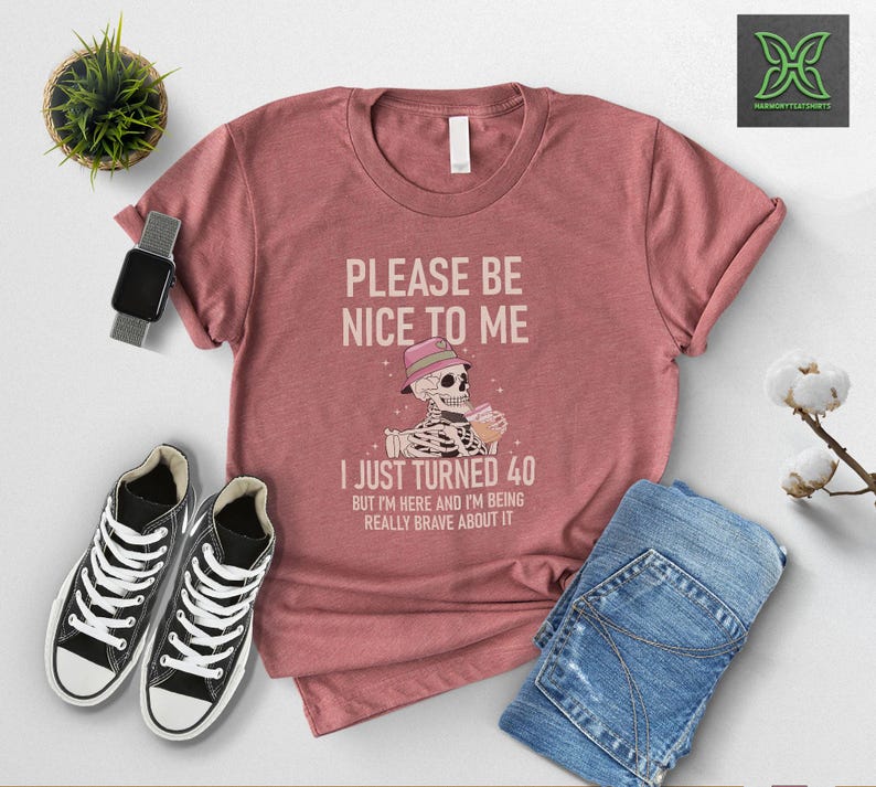 May include: A dusty rose t-shirt with the text "PLEASE BE NICE TO ME I JUST TURNED 40". The graphic shows a skeleton wearing a hat and holding a drink. The shirt is paired with blue jeans and black sneakers.