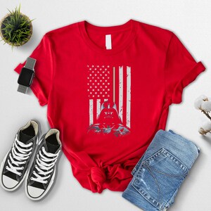 Puede incluir: Camiseta roja con un dise&ntilde;o desgastado de la bandera estadounidense y un gr&aacute;fico de Darth Vader. El dise&ntilde;o fusiona las estrellas y rayas de la bandera con el casco del personaje. La camiseta se muestra con zapatillas negras y vaqueros azules.