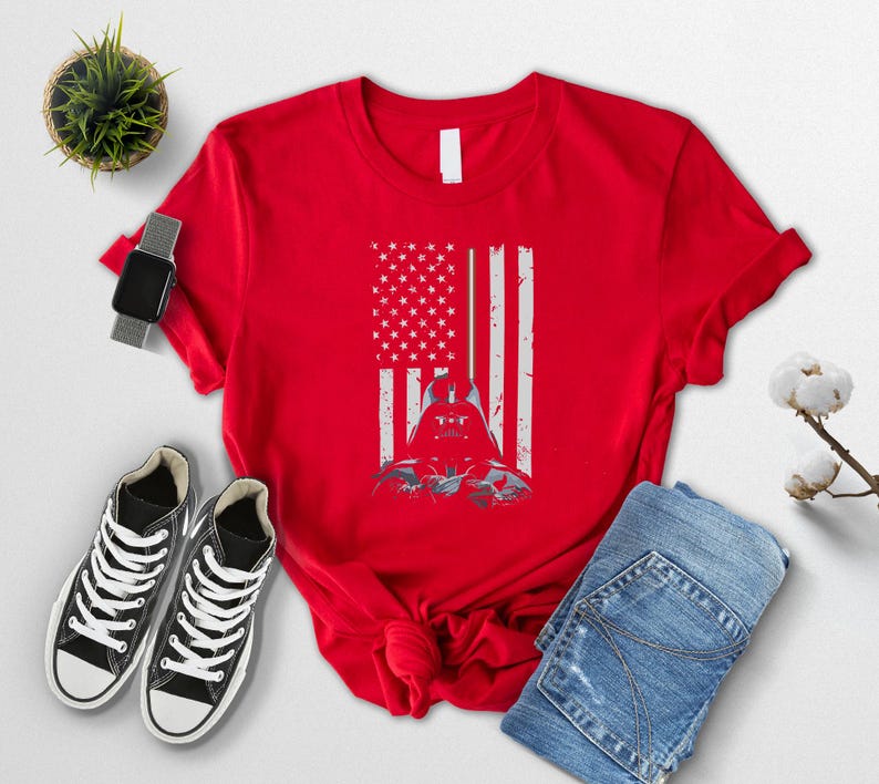 Puede incluir: Camiseta roja con un dise&ntilde;o desgastado de la bandera estadounidense y un gr&aacute;fico de Darth Vader. El dise&ntilde;o fusiona las estrellas y rayas de la bandera con el casco del personaje. La camiseta se muestra con zapatillas negras y vaqueros azules.