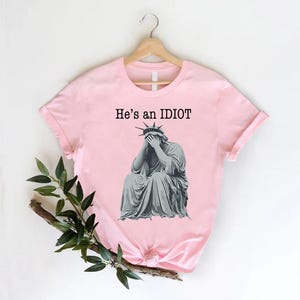 May include: A pale pink t-shirt featuring the text "He's an IDIOT" above a grey illustration of the Statue of Liberty, with her face covered. The t-shirt is displayed on a wooden hanger.