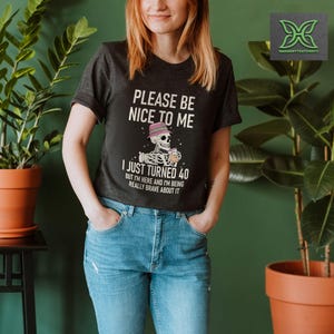 May include: Dark gray t-shirt with the text "PLEASE BE NICE TO ME" and "I JUST TURNED 40". The graphic features a skeleton wearing a hat and holding a drink. The shirt is paired with blue jeans.