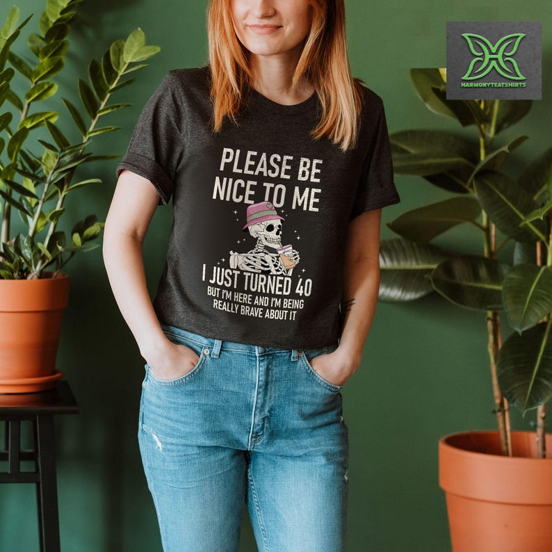 May include: Dark gray t-shirt with the text "PLEASE BE NICE TO ME" and "I JUST TURNED 40". The graphic features a skeleton wearing a hat and holding a drink. The shirt is paired with blue jeans.