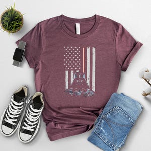 Puede incluir: Camiseta burdeos con un dise&ntilde;o gr&aacute;fico de Darth Vader dentro de una bandera americana desgastada. La bandera tiene rayos de sable l&aacute;ser rojos. La imagen est&aacute; combinada con zapatillas negras y vaqueros azules.