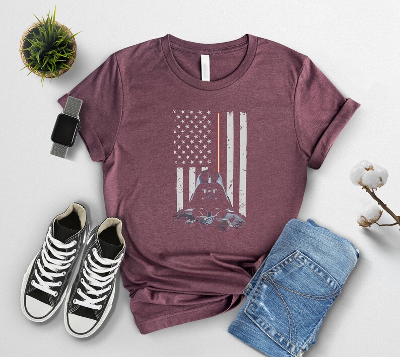 Puede incluir: Camiseta burdeos con un dise&ntilde;o gr&aacute;fico de Darth Vader dentro de una bandera americana desgastada. La bandera tiene rayos de sable l&aacute;ser rojos. La imagen est&aacute; combinada con zapatillas negras y vaqueros azules.