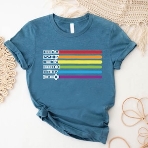 Puede incluir: Una camiseta azul turquesa con un dise&ntilde;o de rayas arco&iacute;ris y siluetas blancas de sables de luz. Los sables de luz est&aacute;n dispuestos verticalmente en l&iacute;nea, con el dise&ntilde;o de rayas arco&iacute;ris detr&aacute;s de ellos.