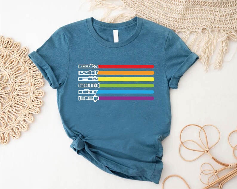 Puede incluir: Una camiseta azul turquesa con un dise&ntilde;o de rayas arco&iacute;ris y siluetas blancas de sables de luz. Los sables de luz est&aacute;n dispuestos verticalmente en l&iacute;nea, con el dise&ntilde;o de rayas arco&iacute;ris detr&aacute;s de ellos.