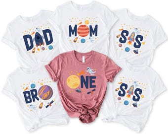 May include: White and pink t-shirts with space-themed designs. The white shirts have the words "DAD", "MOM", "SIS", and "BRO" with rocket ships, planets, and stars. The pink shirt says "ONE" with a planet and astronaut.