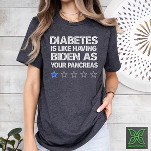 Op de afbeelding: Een donkergrijs T-shirt met witte tekst die "Diabetes is like having Biden as your pancreas." luidt. Onder de tekst staan vijf witte sterren, waarvan de eerste ster blauw is.