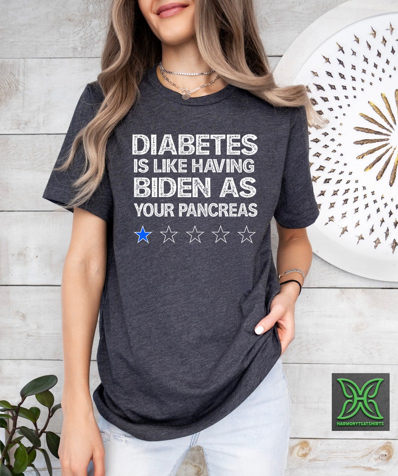 Op de afbeelding: Een donkergrijs T-shirt met witte tekst die "Diabetes is like having Biden as your pancreas." luidt. Onder de tekst staan vijf witte sterren, waarvan de eerste ster blauw is.