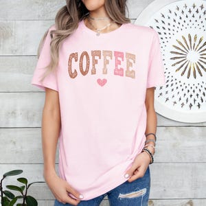 May include: A pink t-shirt with the word "COFFEE" printed in brown, pink, and orange letters with a small pink heart below the word.