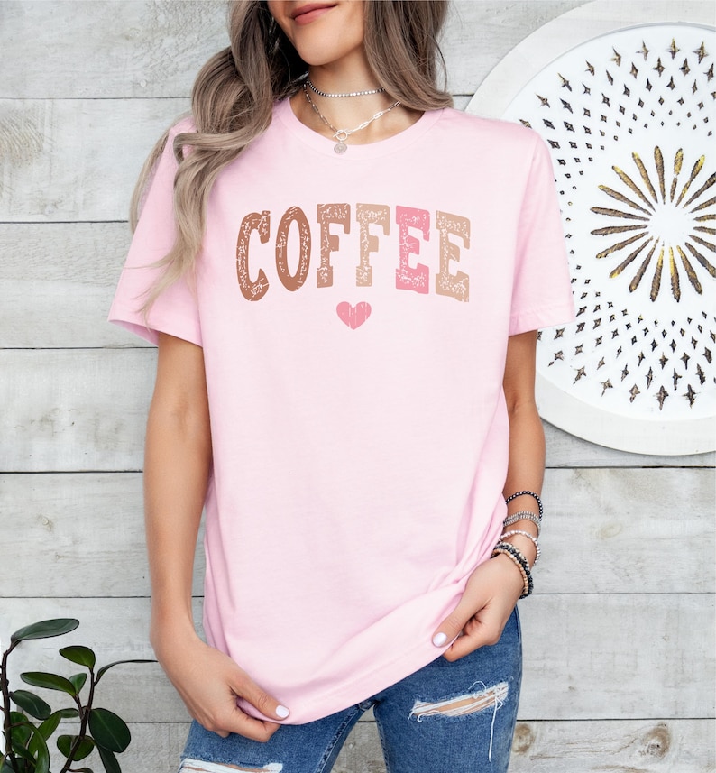 May include: A pink t-shirt with the word "COFFEE" printed in brown, pink, and orange letters with a small pink heart below the word.