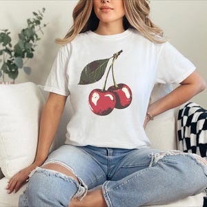 May include: White t-shirt featuring a graphic of two red cherries with a green leaf. The shirt is worn with blue jeans. The cherries have a glossy appearance. The overall theme is casual wear.