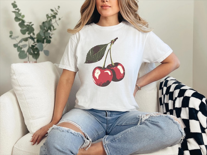 May include: White t-shirt featuring a graphic of two red cherries with a green leaf. The shirt is worn with blue jeans. The cherries have a glossy appearance. The overall theme is casual wear.