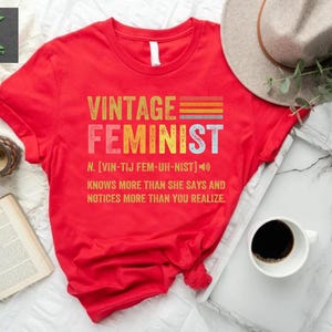 May include: Red t-shirt with a vintage feminist graphic. The graphic features the word "feminist" in a retro font with a rainbow stripe design. The text below defines the word "feminist" as "knows more than she says and notices more than you realize."