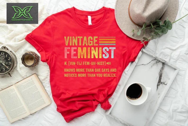 May include: Red t-shirt with a vintage feminist graphic. The graphic features the word "feminist" in a retro font with a rainbow stripe design. The text below defines the word "feminist" as "knows more than she says and notices more than you realize."