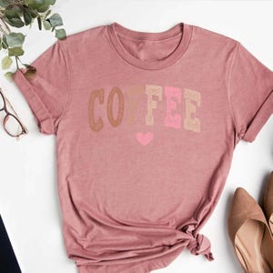 May include: A pink t-shirt with the word "COFFEE" printed in brown, beige, and pink letters. A small pink heart is printed below the word.
