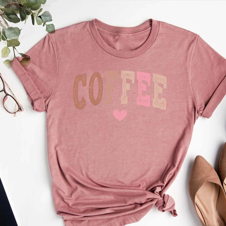 May include: A pink t-shirt with the word "COFFEE" printed in brown, beige, and pink letters. A small pink heart is printed below the word.
