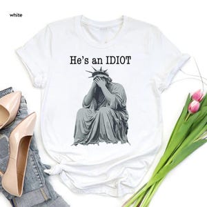 May include: White t-shirt featuring the text "He's an IDIOT" above a grey illustration of the Statue of Liberty, covering her face with her hands. The shirt has a crew neck and short sleeves.