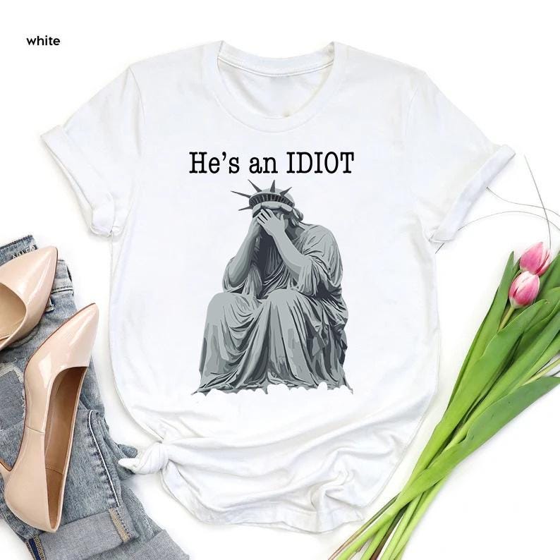 May include: White t-shirt featuring the text "He's an IDIOT" above a grey illustration of the Statue of Liberty, covering her face with her hands. The shirt has a crew neck and short sleeves.