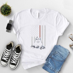Puede incluir: Camiseta blanca con un dise&ntilde;o de Darth Vader y un sable de luz, sobre un dise&ntilde;o estilizado de la bandera estadounidense. La camiseta se combina con vaqueros azules y zapatillas negras. Tambi&eacute;n se ven un reloj y una peque&ntilde;a planta.