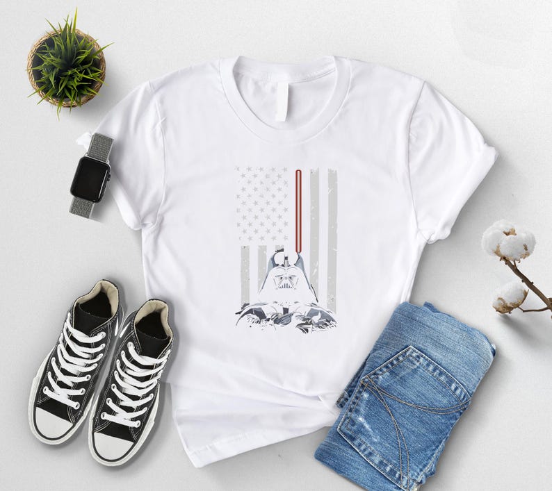 Puede incluir: Camiseta blanca con un dise&ntilde;o de Darth Vader y un sable de luz, sobre un dise&ntilde;o estilizado de la bandera estadounidense. La camiseta se combina con vaqueros azules y zapatillas negras. Tambi&eacute;n se ven un reloj y una peque&ntilde;a planta.