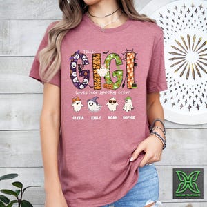 Op de afbeelding: Een mauvekleurig T-shirt met de tekst "This Gigi loves her spooky crew" in een Halloween-ontwerp. Het ontwerp bevat geesten, pompoenen en de namen Olivia, Emily, Noah en Sophie.