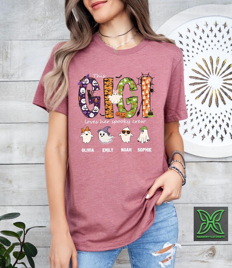 Op de afbeelding: Een mauvekleurig T-shirt met de tekst "This Gigi loves her spooky crew" in een Halloween-ontwerp. Het ontwerp bevat geesten, pompoenen en de namen Olivia, Emily, Noah en Sophie.