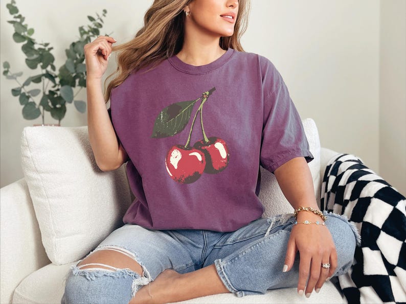 May include: A purple t-shirt featuring a distressed graphic of two cherries with a green leaf. The shirt is a solid color and has a relaxed fit. The model is wearing blue jeans.