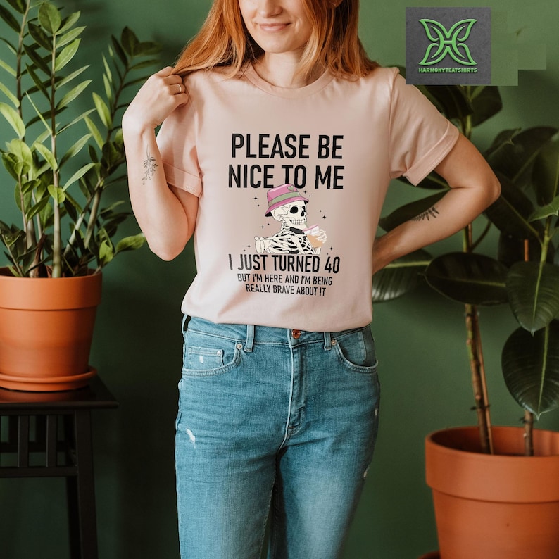 May include: A light pink t-shirt with the text "PLEASE BE NICE TO ME" and "I JUST TURNED 40". The graphic features a skeleton wearing a hat and holding a drink. The shirt is paired with blue jeans.